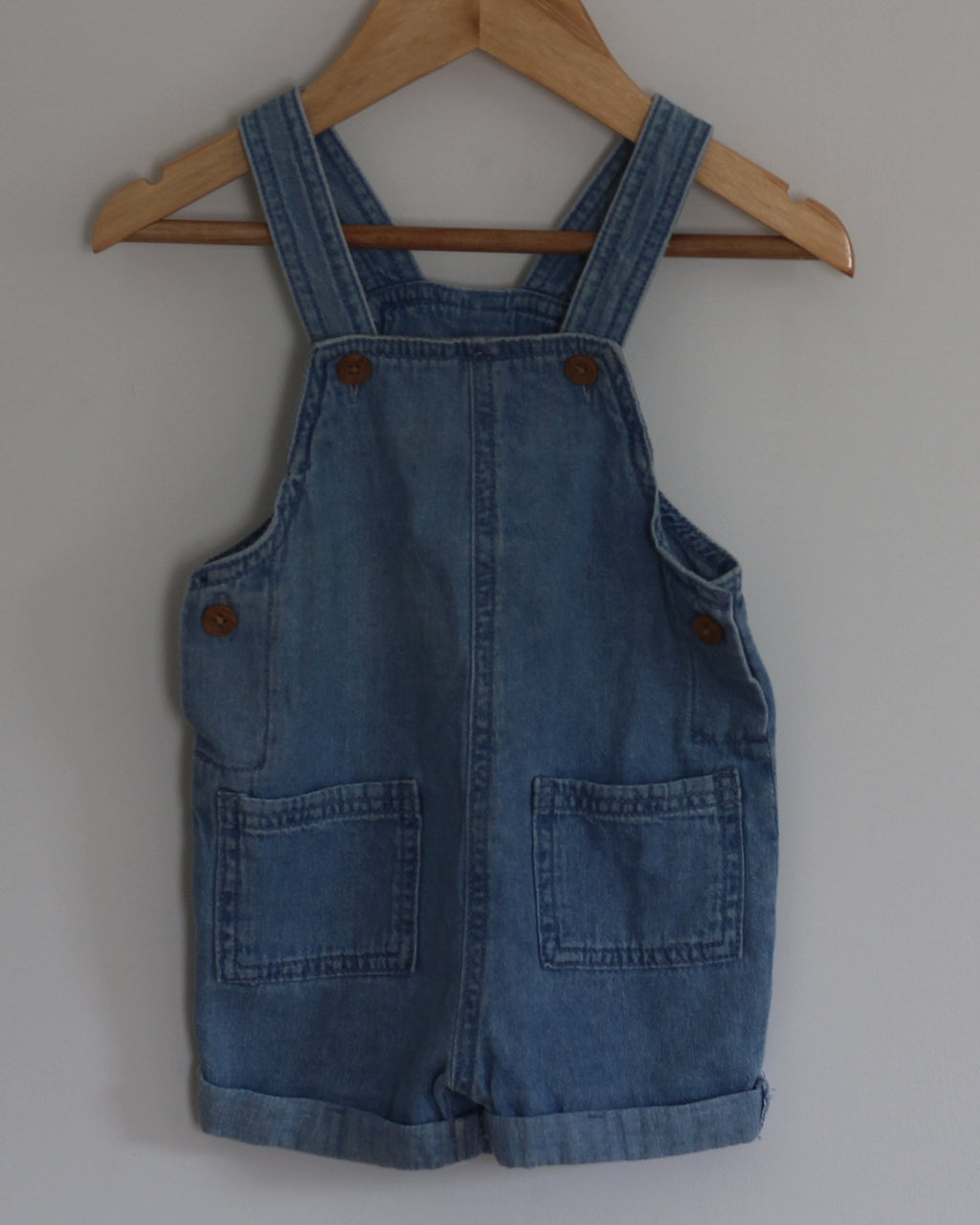 A blue denim baby overall romper hangs from a wooden hanger on a wall hook against a plain light gray wall.