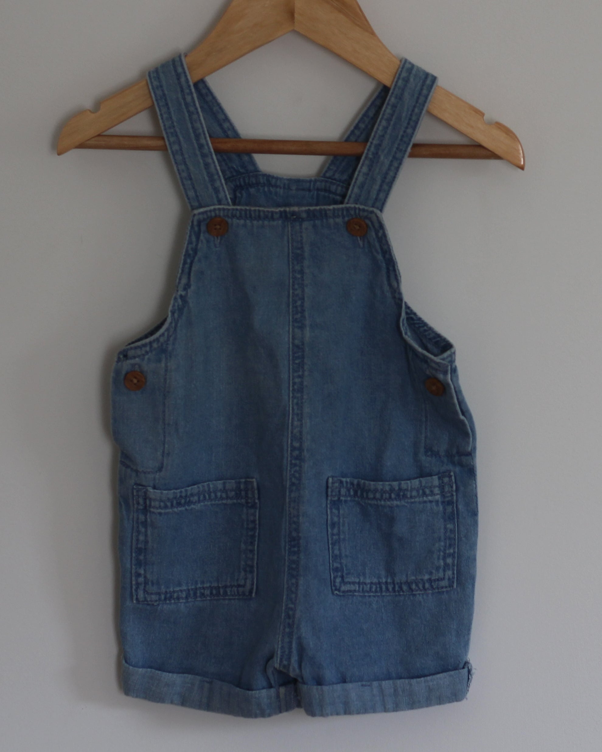 A blue denim baby overall romper hangs from a wooden hanger on a wall hook against a plain light gray wall.