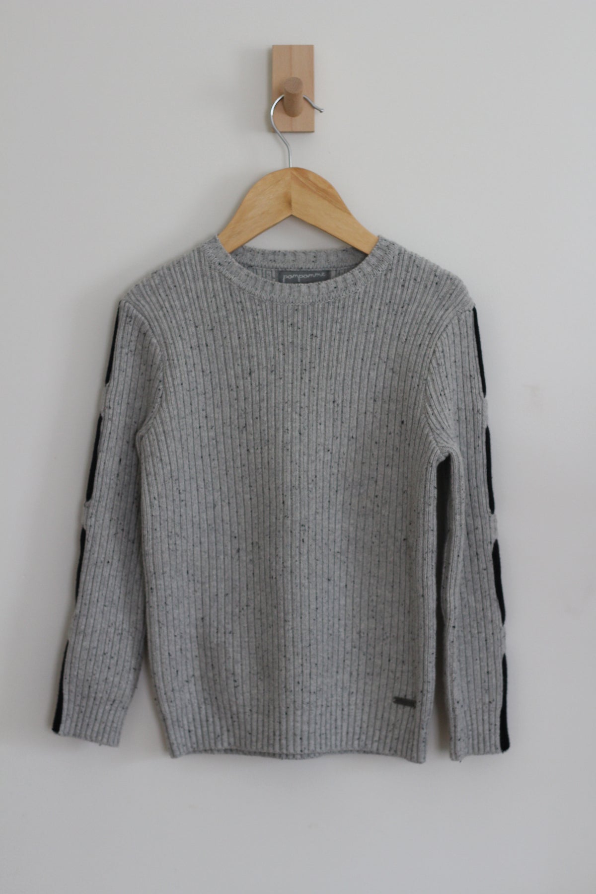 A gray ribbed knit sweater with black stripe details on the sleeves, hanging on a wooden hanger from a wall hook.