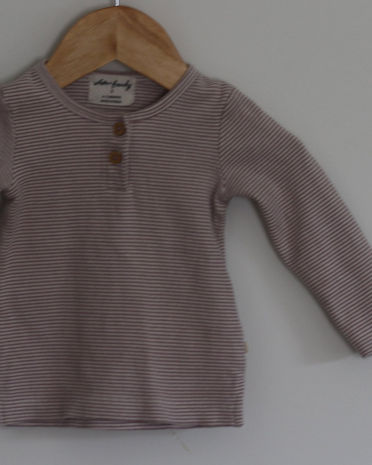 A long-sleeve baby henley shirt in a muted dusty mauve color, hanging on a wooden hanger from a wall peg against a plain light gray wall.